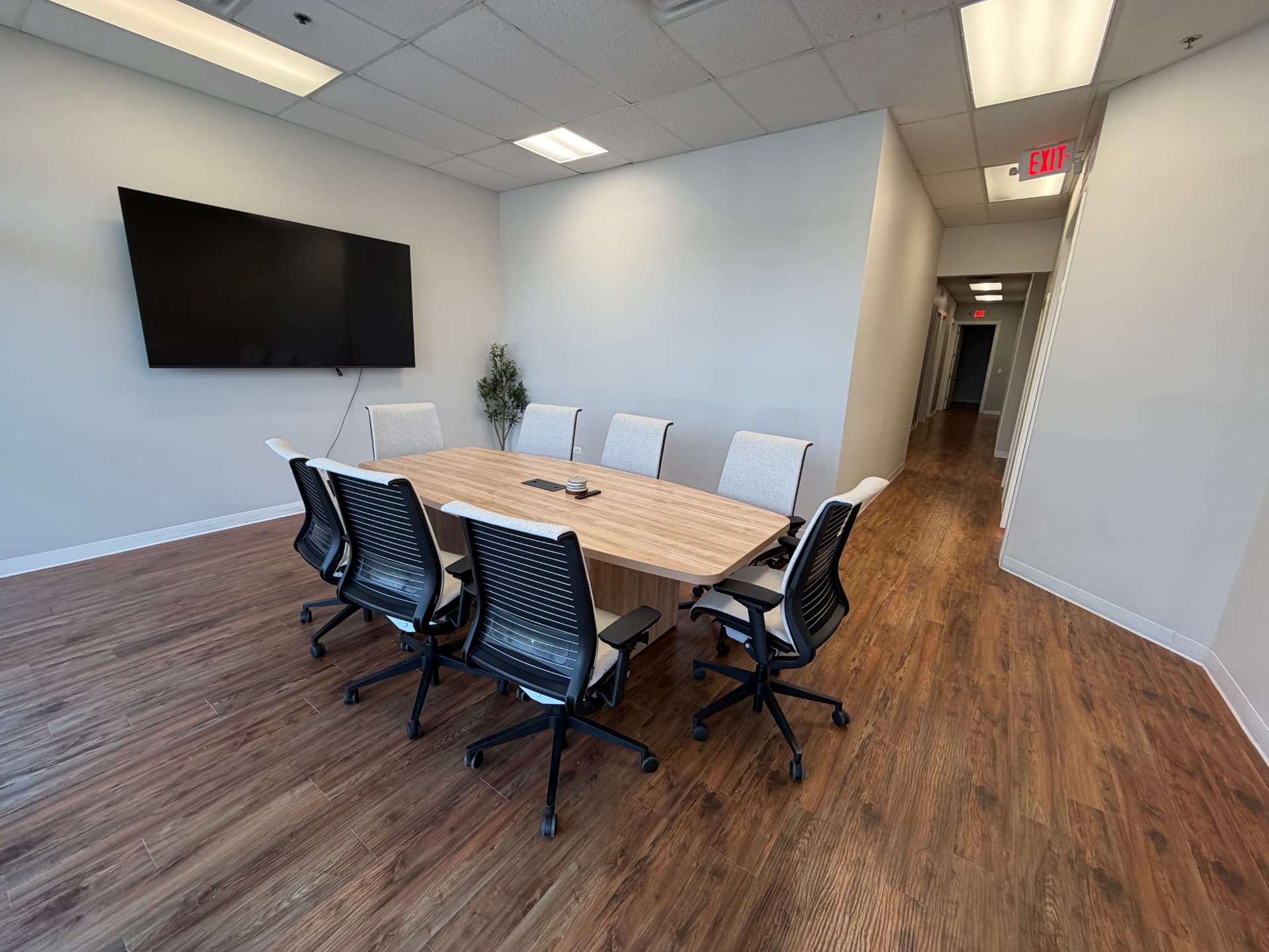 Shared conference room with an eight-seat table, wall-mounted screen and a hallway beyond.